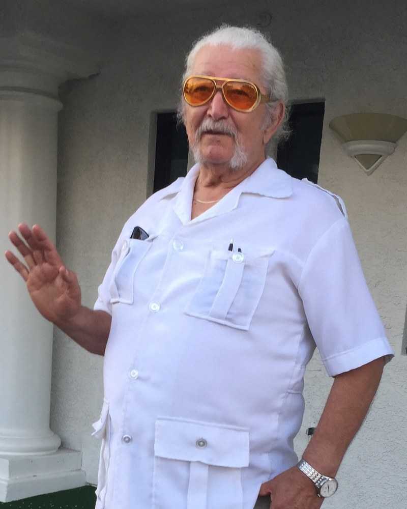 Tata outdoors in amber-gold aviator sunglasses and a white guayabera shirt, one hand raised in a wave.