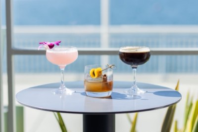 Three cocktails on a small table with the bay blurred behind them.
