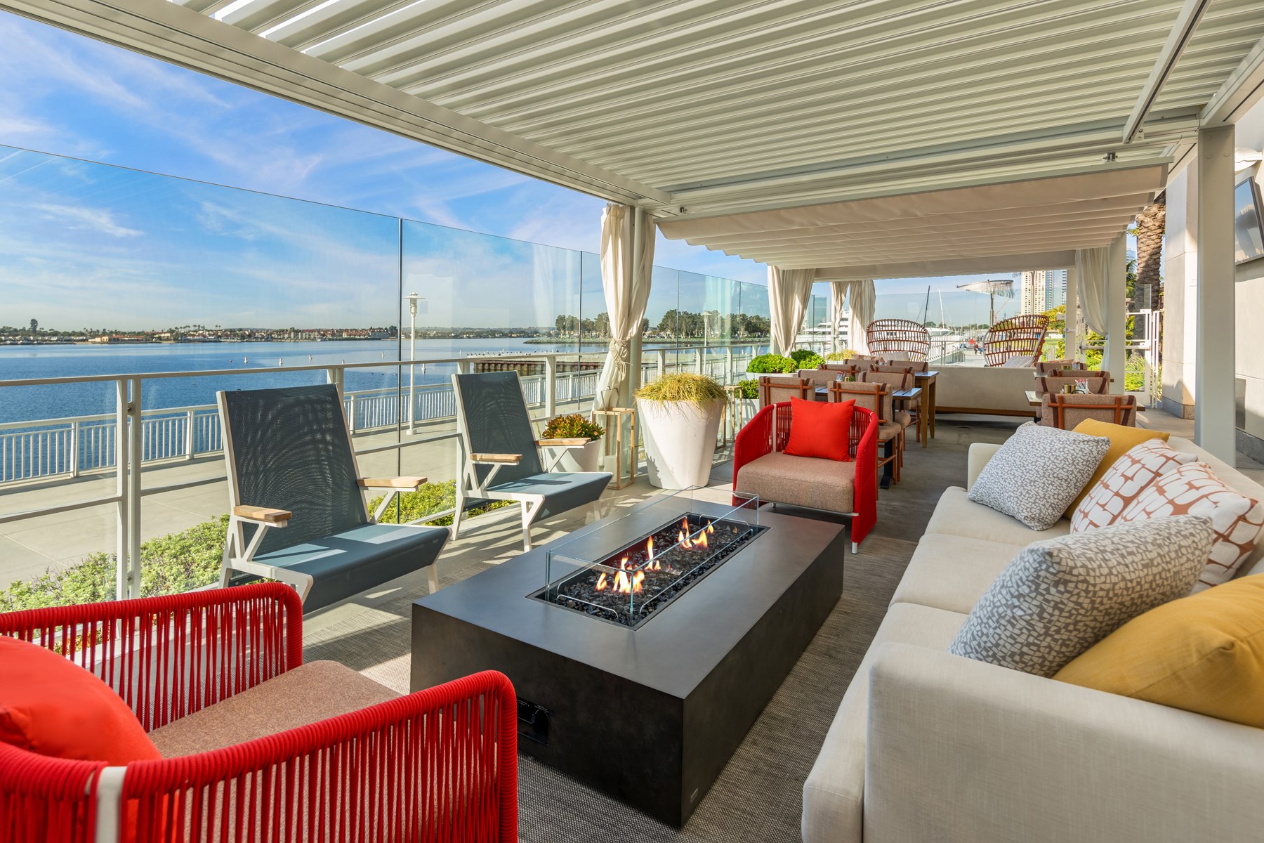 The open-air waterfront patio at Hudson and Nash: lounge seating and a long fire pit, glass railing, and San Diego Bay beyond.